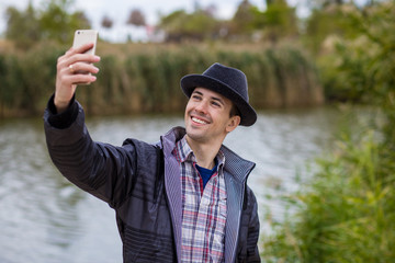 Obraz premium Young happy man taking selfie in fashionable clothes on the river bank 