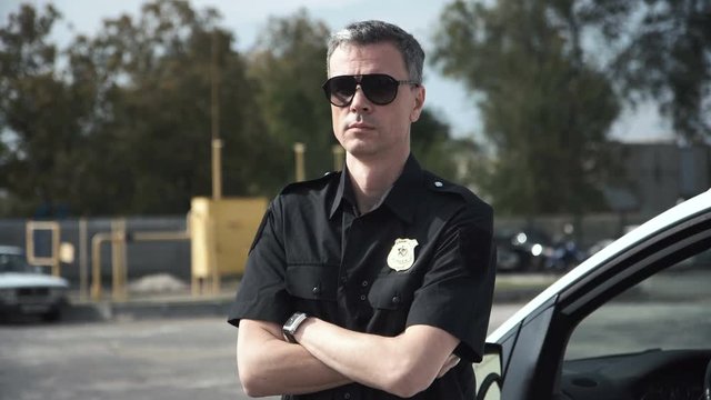 Serious-looking police officer with badge standing near his car and looking at camera.