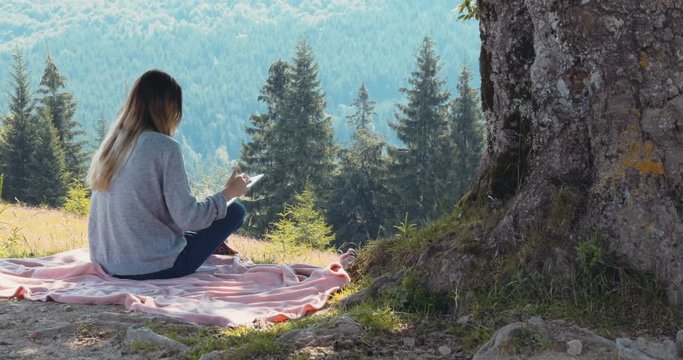 Sunshine top mountain back view young woman sitting grass blanket hillside summer enjoying beautiful landscape forest nature using digital tablet tapping touchscreen traveling trendy technology online