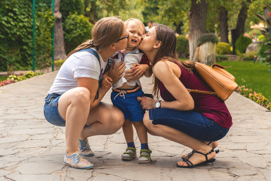 Two girls (mom) kiss their capricious little boy child in the park. Not a traditional family.