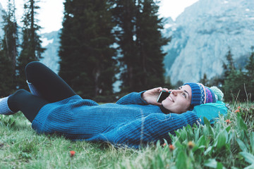 Young beautiful traveler woman with backpack, hat lying in the grass and flowers and chating on mobile phone, smilling in the stunning mountain wilderness in front of amazing cold lake after  hiking