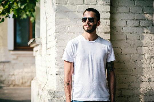 Young stylish man wearing white blank t-shirt with beard in glasses, standing on the street on city background. Street photo