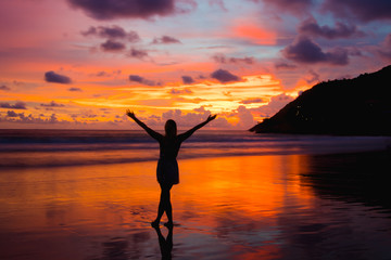 Silhouette of woman with hands up while standing on the sea beach at sunset.