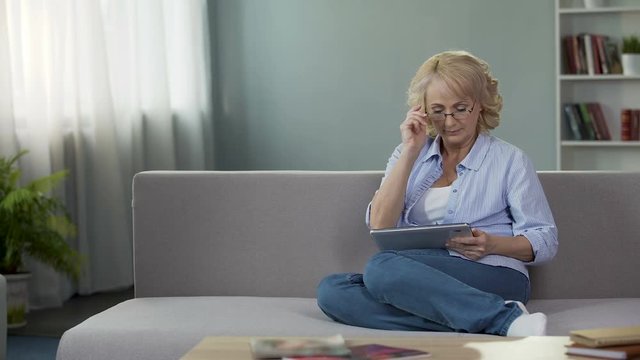 Adult woman sitting on sofa and holding tablet, buying new eyeglasses online