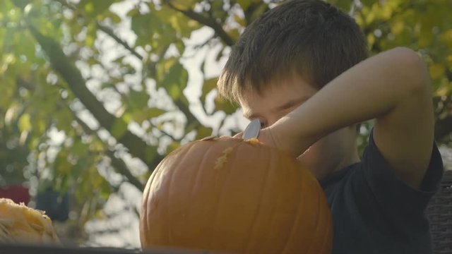 Little boy strokes spoonful Halloween creepy head. Child hands carve halloween pumpkin scarecrow head. Preparing Halloween Pumpkins. Slow motion shot of autumn garden.