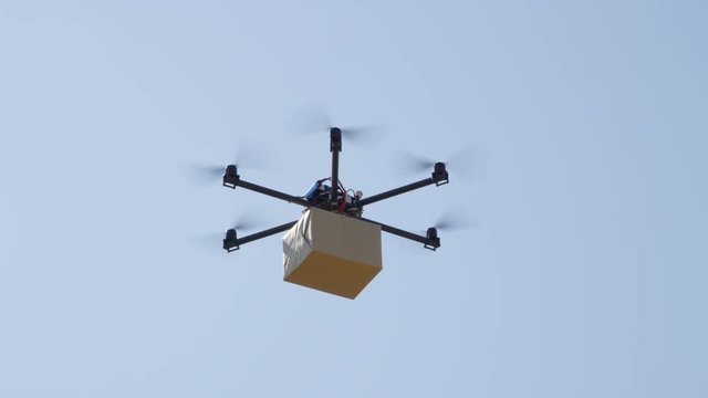 CLOSE UP UAV drone delivery. Multicopter flying big brown package over blue sky. Drone delivering post package to your home. Futuristic shipment by helicopter drone. Multirotor logistics and transport