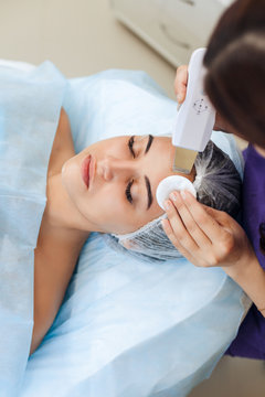 The doctor-cosmetologist makes the apparatus a procedure of ultrasound cleaning of the facial skin of a beautiful, young woman in a beauty salon. Cosmetology and professional skin care.