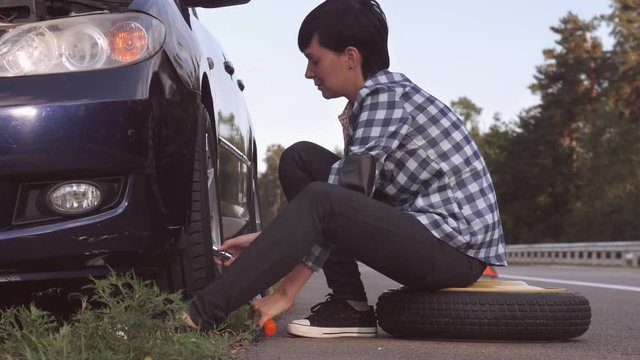 Caucasian young woman wearing casual shirt and jeans changing a tire sitting on stepney near car on the roadside. Brunette traveller outdoors