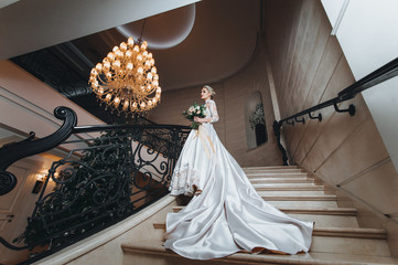 Beautiful young woman in a white dress with long train is climbs up the stairs in a classic...