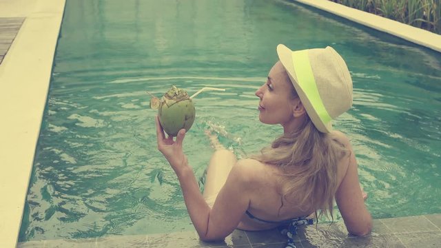 The young slender woman in Bikin drinks coconut milk from a coco on the edge of the pool in the tropical resort,retro effect