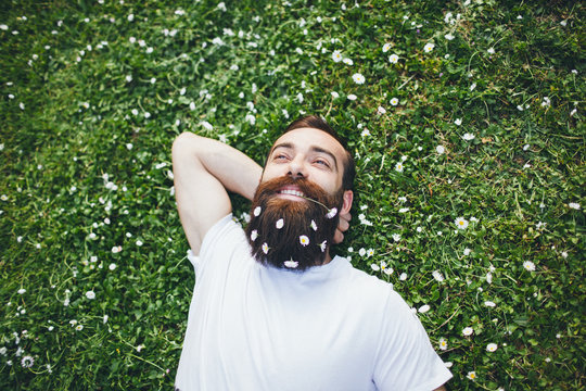Smiling young man relaxing on grass with flowers in his beard