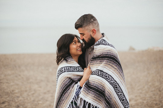 Couple wrapped in Mexican blanket and hugging and smiling on beach