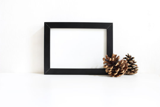 Black blank wooden frame mockup with pine cones lying on the white table. Poster product design. Styled stock feminine photography. Home decor. Christmas winter concept.