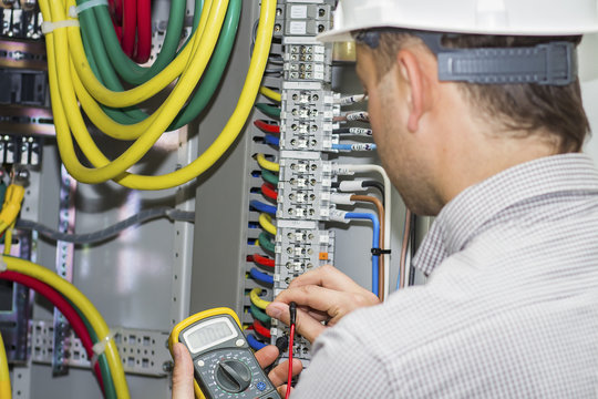 Engineer tests supply circuit in powerful panel of distribution cables on terminal box background. Electrician with multimeter measures voltage in fuse box