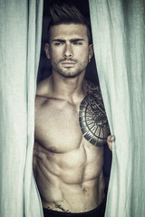 Portrait of sexy shirtless muscular man next to window curtains during the day, wearing only jeans