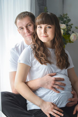 Young pregnant woman and her husband are hugging, touching the belly near window smiling and looking at camera