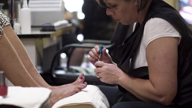 Beautician focusses on painting the toenails red of a woman in a pedicure chair. Slider moves camera from left to right.