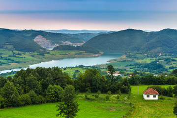 A small white house with a brown roof on the background of the coastal valley of the lake with large hills in cloudy weather. Rovni lake, Valjevo, Serbia.
