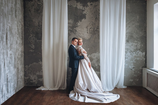 Beautiful happy young newlyweds gentle embrace in a gray studio. White decorative drape on an abstract background. 