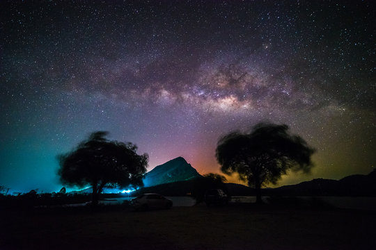 Panoramic milky way galaxy with foreground of mountain, colorful city light and silhouette trees at Lam Isu Reservoir, Kanchanaburi province, Thailand. Long exposure, low light.