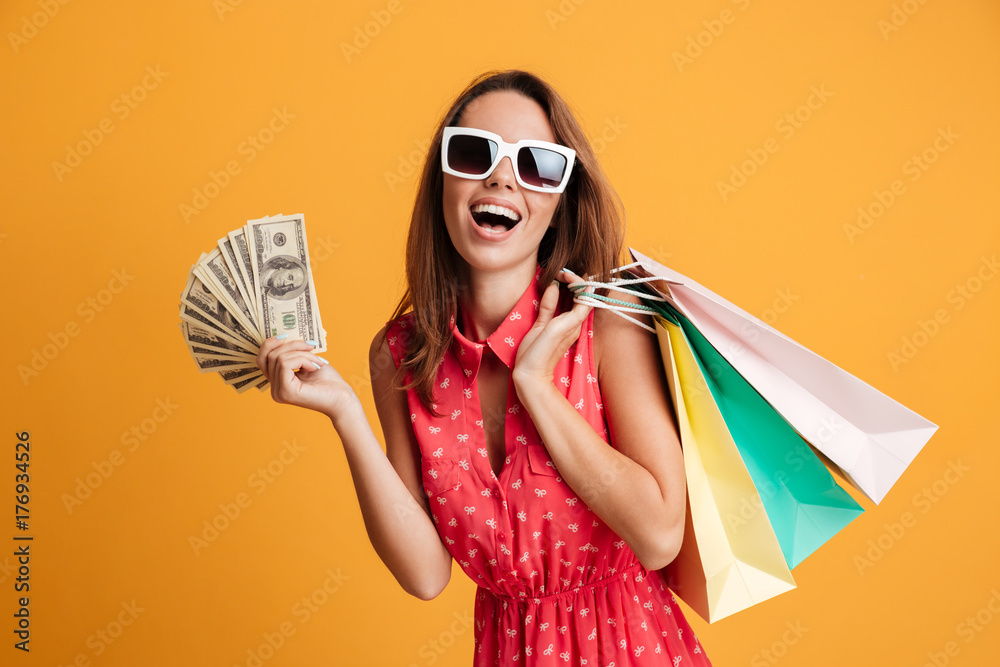 Sticker photo of happy young woman in sunglasses holding fan of money and colorful shopping bags, looking at