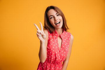 Close-up portrait of funny brunette girl showing peace gesture, looking at camera with open mouth