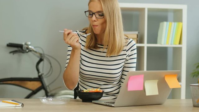 Attractive busy businesswoman in glasses eating salad in the office during lunch time.