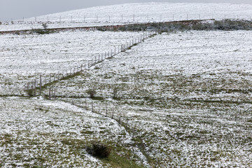 A fence running up on a mountain, in the midst of snow covering partially the grass meadows