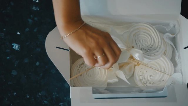 The confectioner puts the meringue in a box on a stick with a gentle bow. Female hands with a white bracelet of beads neatly places in the empty cardboard box marshmallows on a thin long straw. After