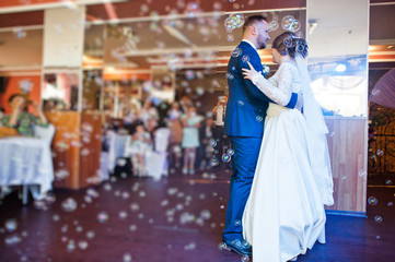Obraz premium Beautiful wedding couple performing their first dance in the restaurant with different lights and bubbles and guests on the background.