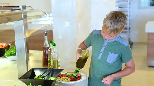 Little child takes food in self-service restaurant. Kid pouring some olive oil from bottle on sausages, vegetables and omelette on white plate. Real time full hd video footage.