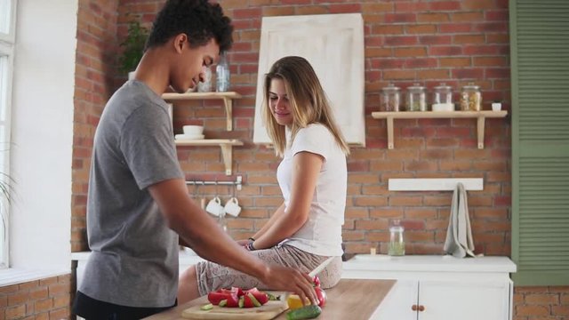 Handsome african guy preparing salad, cutting lemon and squeezing juice from it in a funny way while his caucasian girlfriend is sitting on the table in the kitchen and laughing