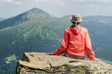 Young woman in a red jacket sitting on top of a mountain enjoying a spectacular view of the mountain landscapes. © Ant