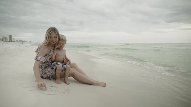 Young woman and child sitting on ocean shore. Female in hat and little boy sat comfortably on waterfront on warm summer evening. Nicely about something babbling mom shows his finger to son on floating