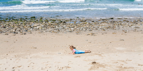 Fototapeta na wymiar Little girl of elementary age laying on sandy beach with ocean water in background. Kid playing in sand. Childhood summer vacation concept.