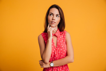 Photo of thinking young woman in red dress touching her chin, looking upward, isolated on yellow background