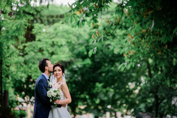 Fototapeta premium Handsome groom and pretty bride with white bouquet stand on green lawn