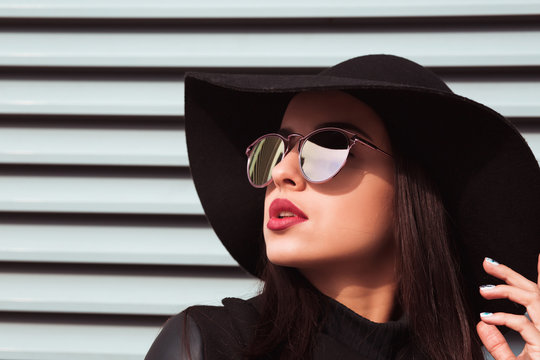 Closeup portrait of young fashionable model wears hat and sunglasses. Woman posing at the background of shutters in sunny day