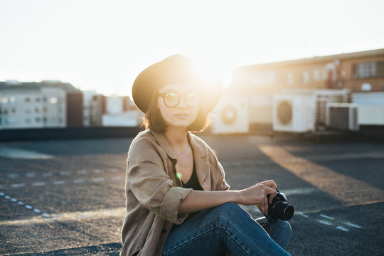 Smiling and laughing young model teenager or woman in hipster outfit, glasses and fedora hat holds vintage analog photo camera, sits on top of rooftop during summer sunset