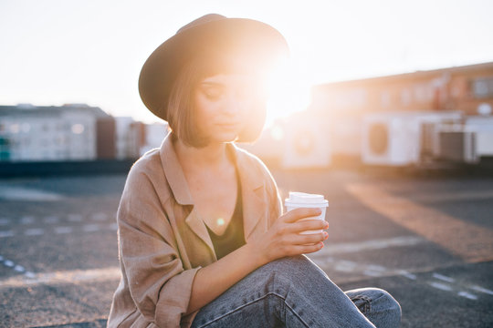 Attractive young millennial student or fashion model in hipster outfit poses for camera on top of rooftop with sun light leaks, smiles and holds coffee cup to go