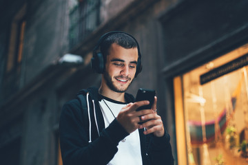 Handsome young man in black hoodie wears big wireless music headphones and changes audio tracks on smartphone, he smiles when looks at phone screen