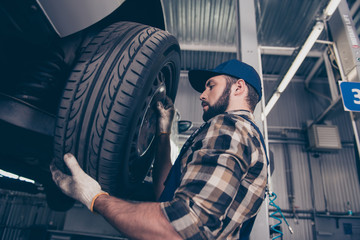 Fototapeta na wymiar Low angle side profile shot of expert specialist technician changes tires, tyres of lifted up car, at auto workshop, wears checkered shirt, uniform costume, hat headwear