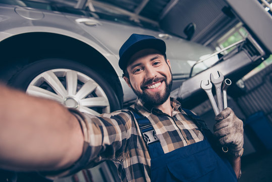Attractive brunet bearded automotive expert with beaming smile makesphoto with metal mechanical keys in arm, in special safety outfit uniform, checkered shirt, hat head lifted up vehicle behind