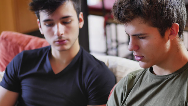 Two Young Male Friends Talking And Chatting While Sitting On Sofa At Home, Relaxing With Serious Expression