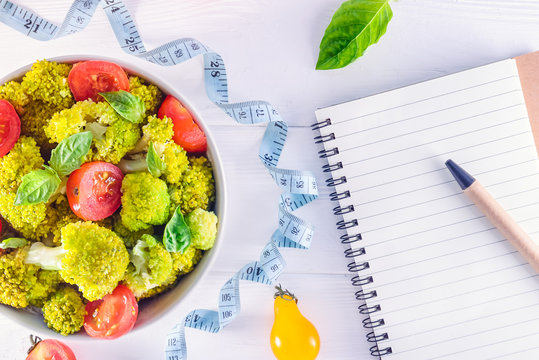 Top view Salad Bowl with cherry tomatoes, broccoli, measuring tape and diet book on the white wooden background. Healthy lifestyle concept. Detox, diet, vegitarian. Selective focus. Text space.