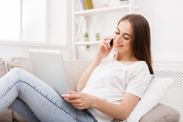 Fototapeta premium Young woman talking on mobile and using laptop at home on the couch.