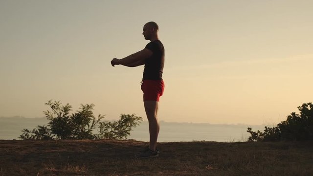 side view man with short hair wearing sport clothes doing sit-ups working out keeping hands in front of him on the edge of hill at sunset time slow motion