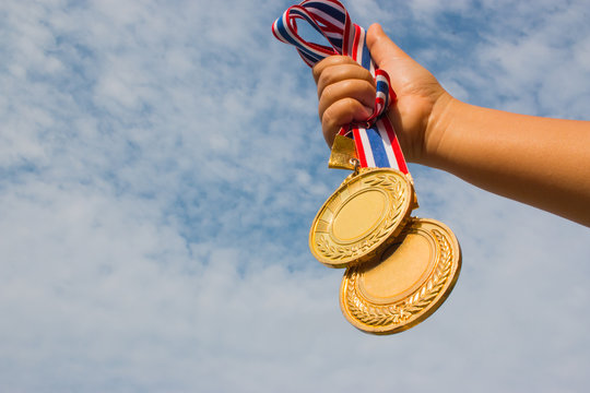 Winner hand raised and holding two gold medals with Thai ribbon against blue sky. Golden medal is medal awarded for highest achievement for sport or business. Success Awards concept