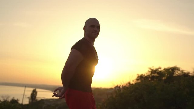 slow motion portrait of strong caucasian bald bodybuilder man wearing black T-shirt posing smiling on camera showing arm muscles with summer sunset over sea background
