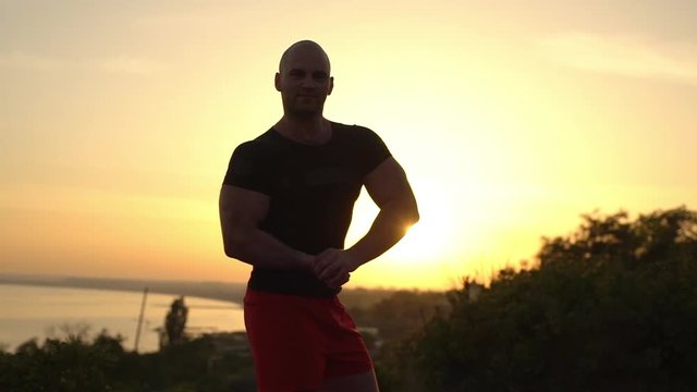 footage of muscular bald bodybuilder man wearing black T-shirt posing smiling on camera showing biceps with summer sunrise over sea background slow motion 